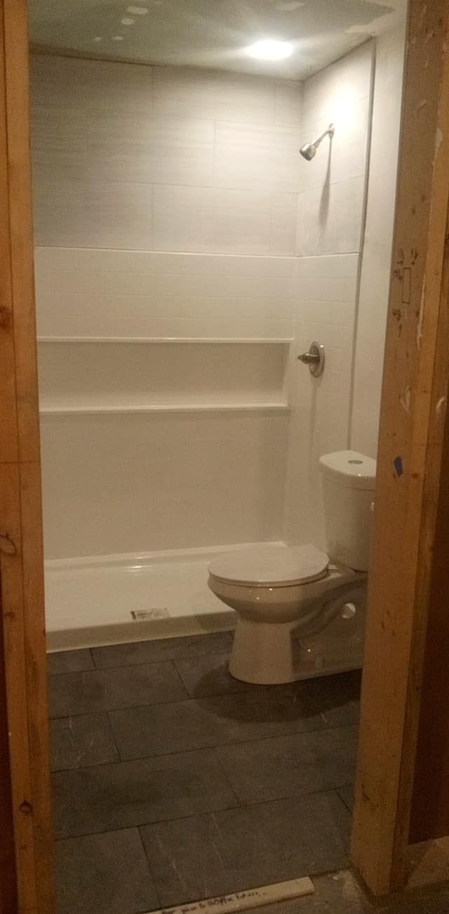 Bathroom interior with a white shower, toilet, and gray flooring. The room is framed by unfinished wooden supports.