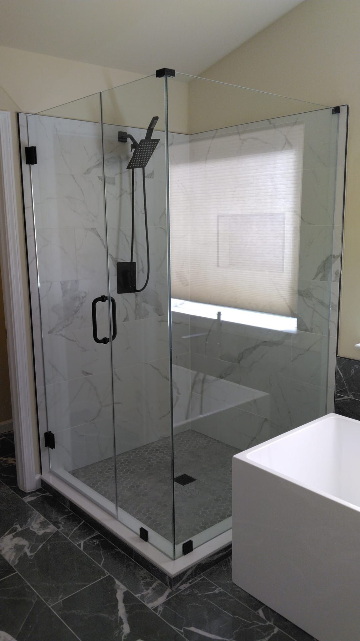 Modern glass shower enclosure with black hardware, marble-look tile, and a white base, next to a window with a shade.