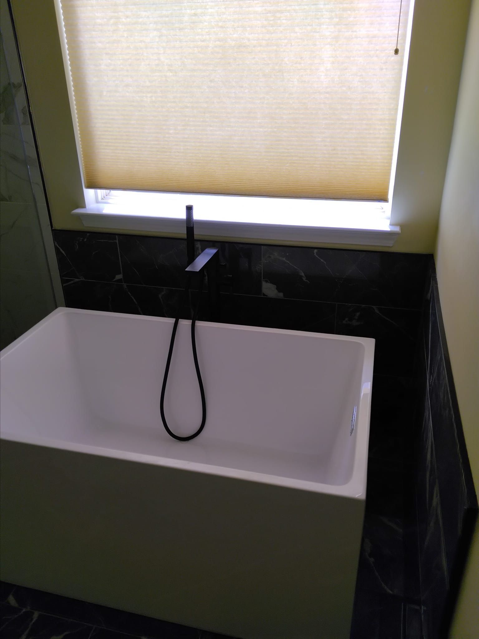 White bathtub with black faucet, below window with beige shade, set in bathroom with black tile and yellow walls.