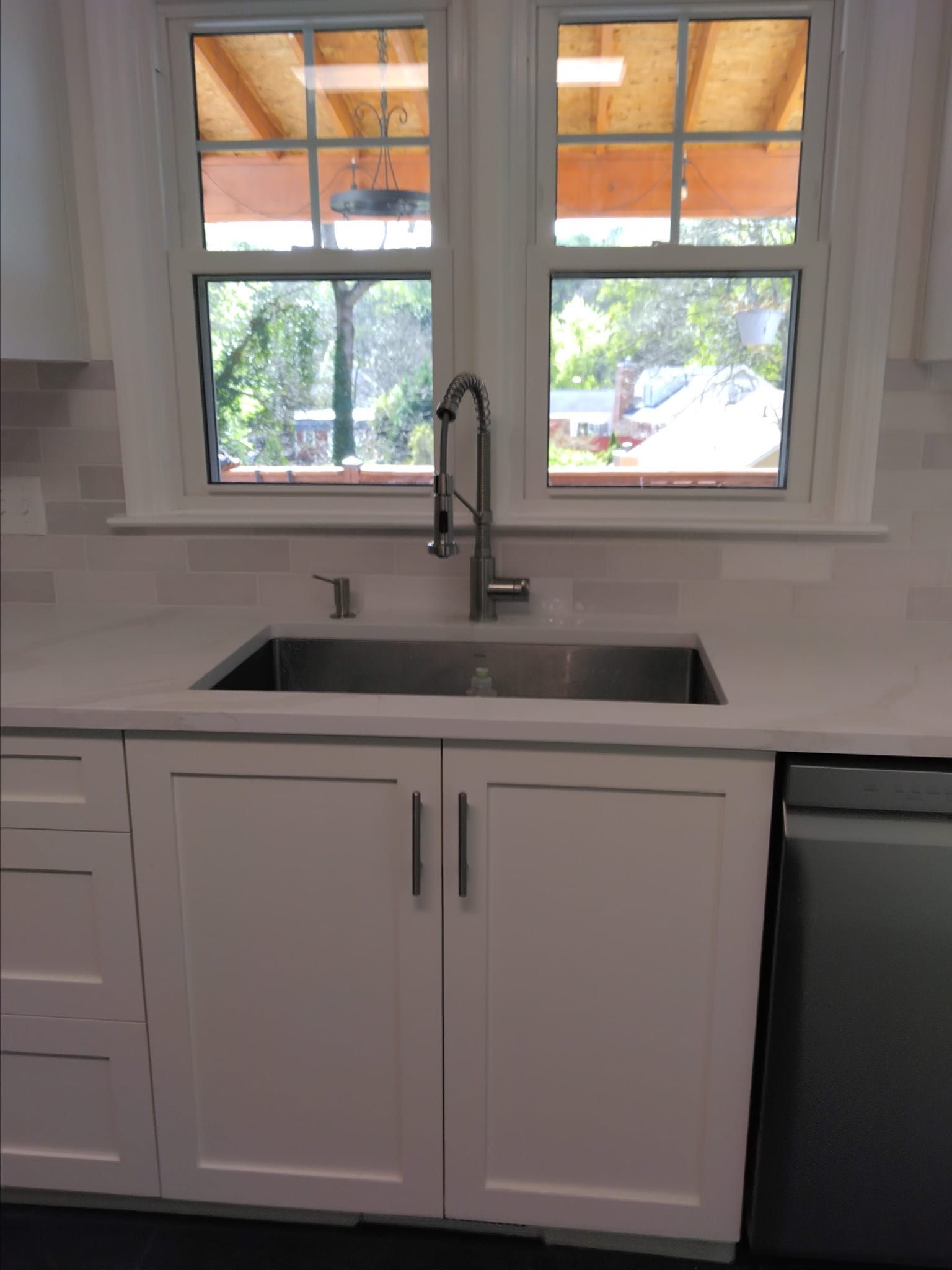 Stainless steel kitchen sink under a window with a faucet, white cabinets, and a dishwasher.