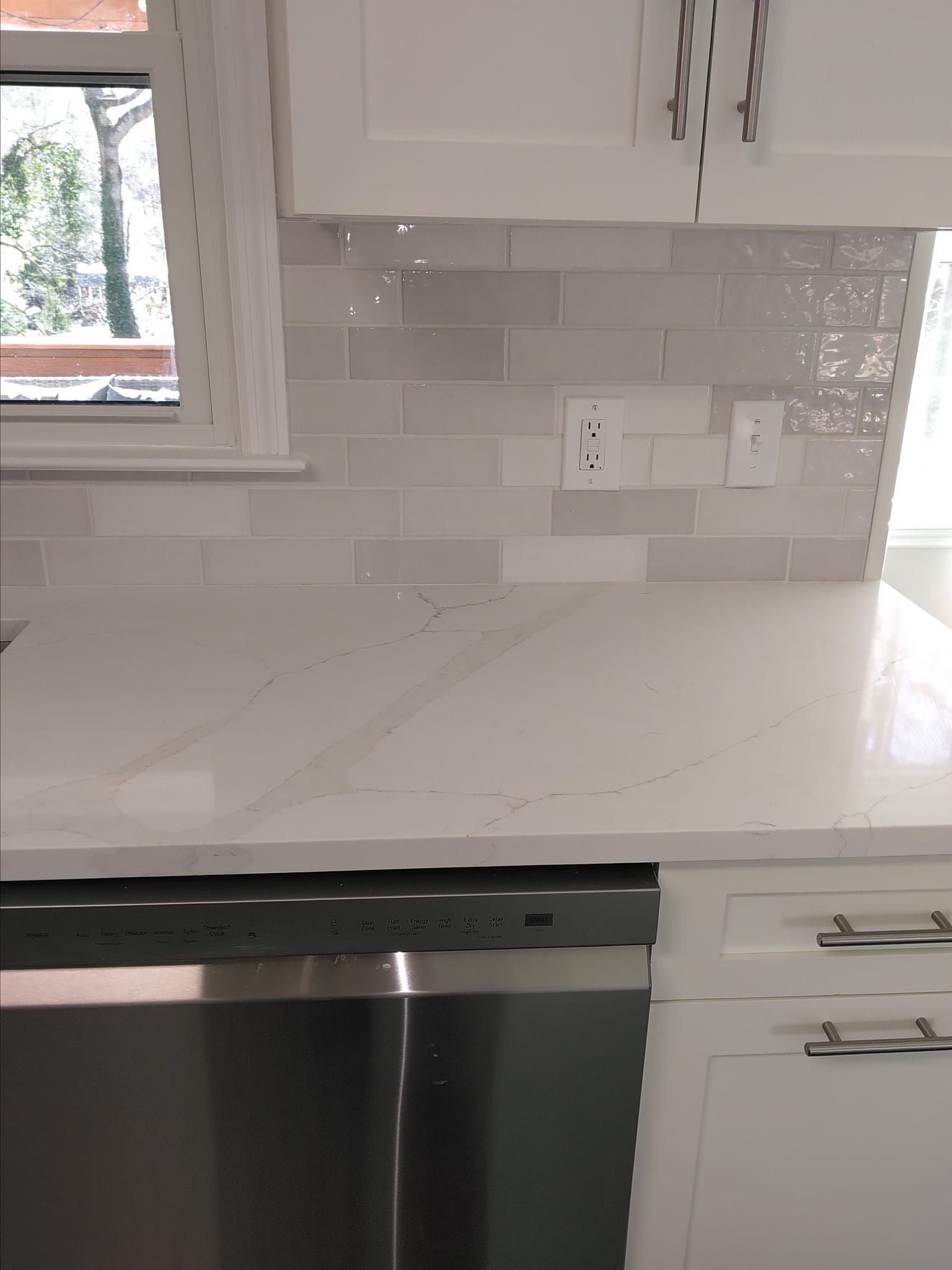 White kitchen with light gray tiled backsplash, white countertops, and stainless steel dishwasher.