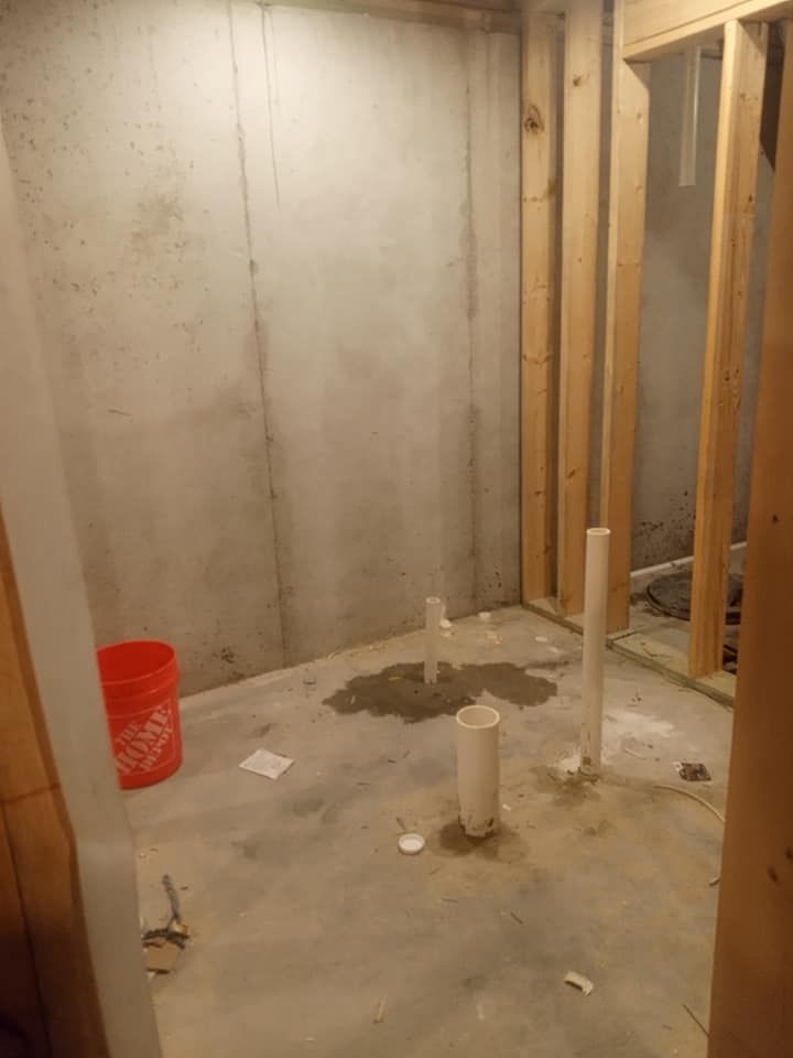Basement bathroom framing with exposed concrete wall, plumbing, bucket, and framing studs.
