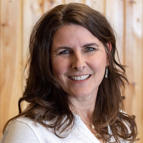 A woman with long brown hair is smiling in front of a wooden wall.