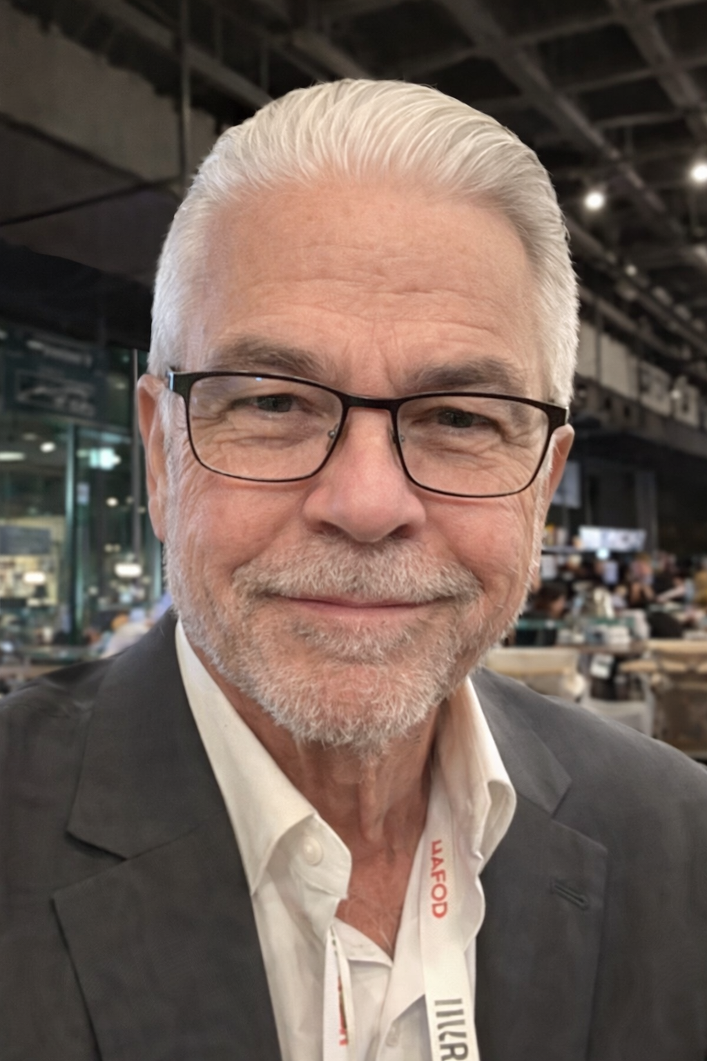 A man with white hair and glasses smiles in a dimly lit, professional setting, wearing a blazer and collared shirt.