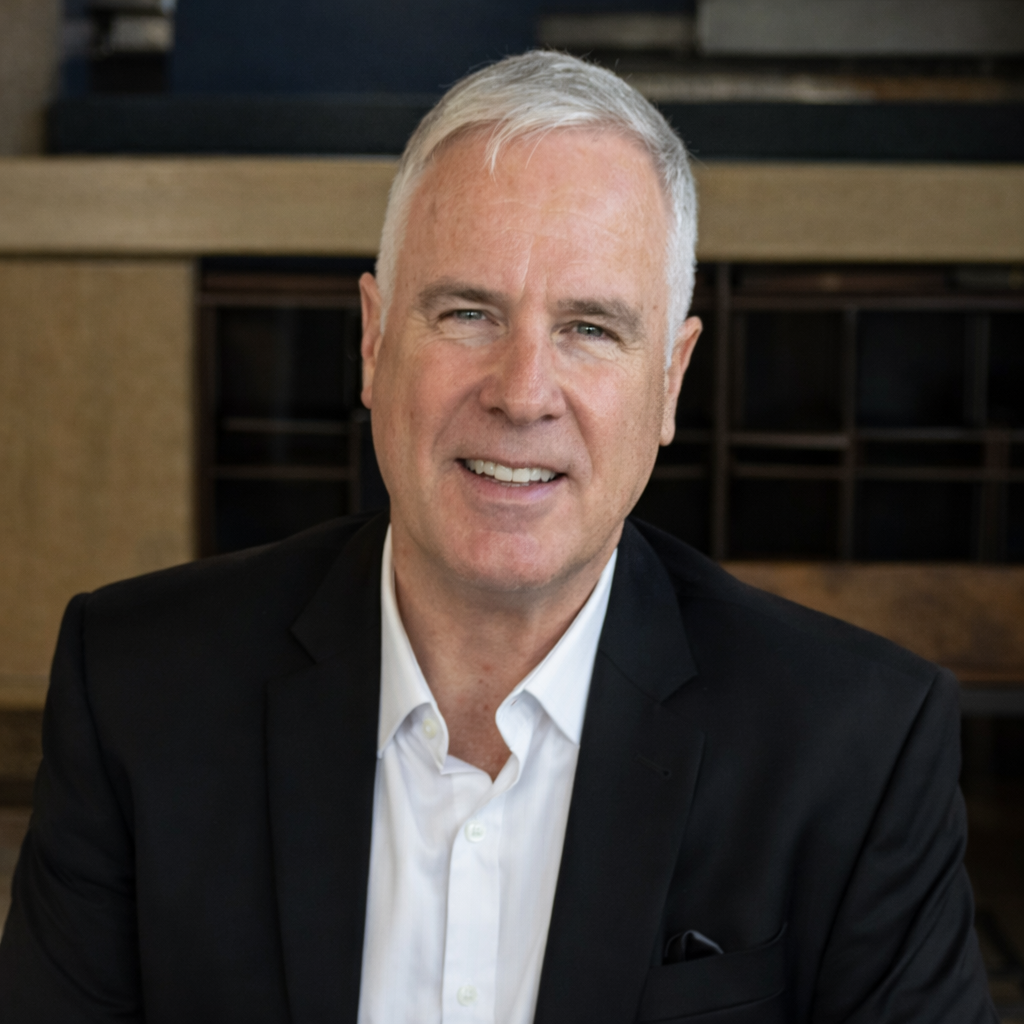 A person with gray hair wearing a black blazer and white collared shirt, smiling against a dark, indoor background.