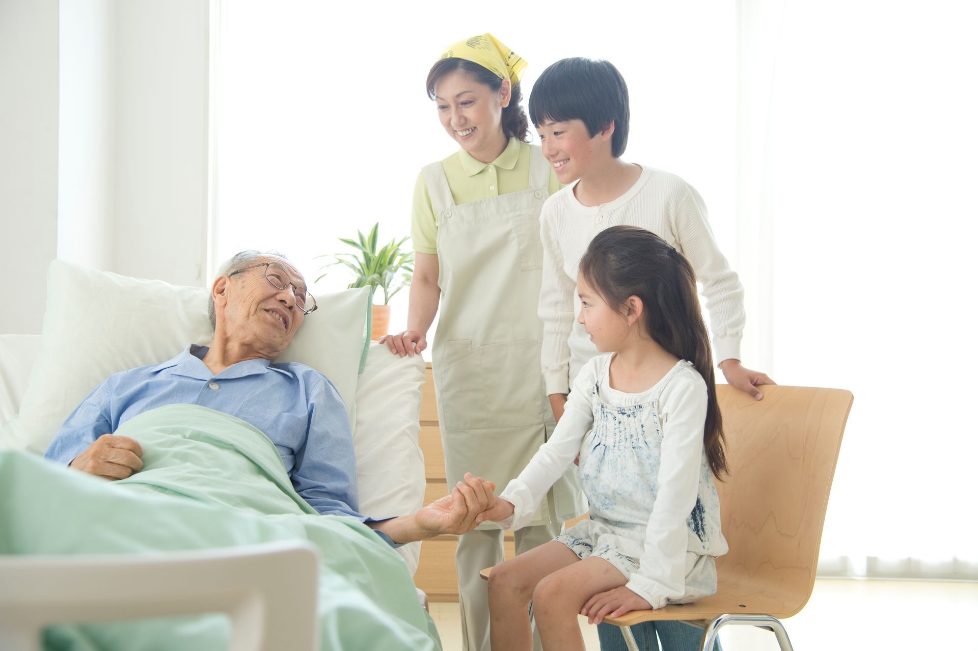 Family visiting a senior lying in bed; they are smiling and holding hands. Light, bright room.