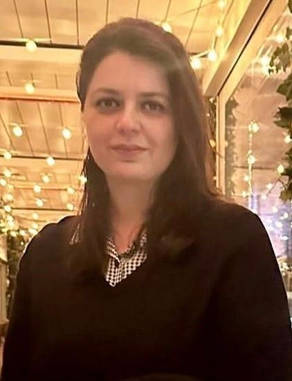 Woman with dark hair wearing a black sweater, small patterned top. Sitting indoors, with string lights in the background.