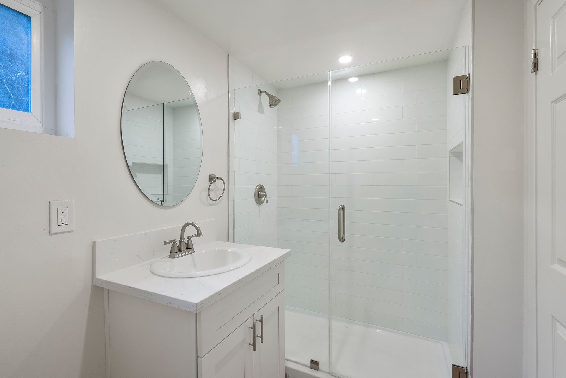 White bathroom with vanity, oval mirror, shower, and small window.