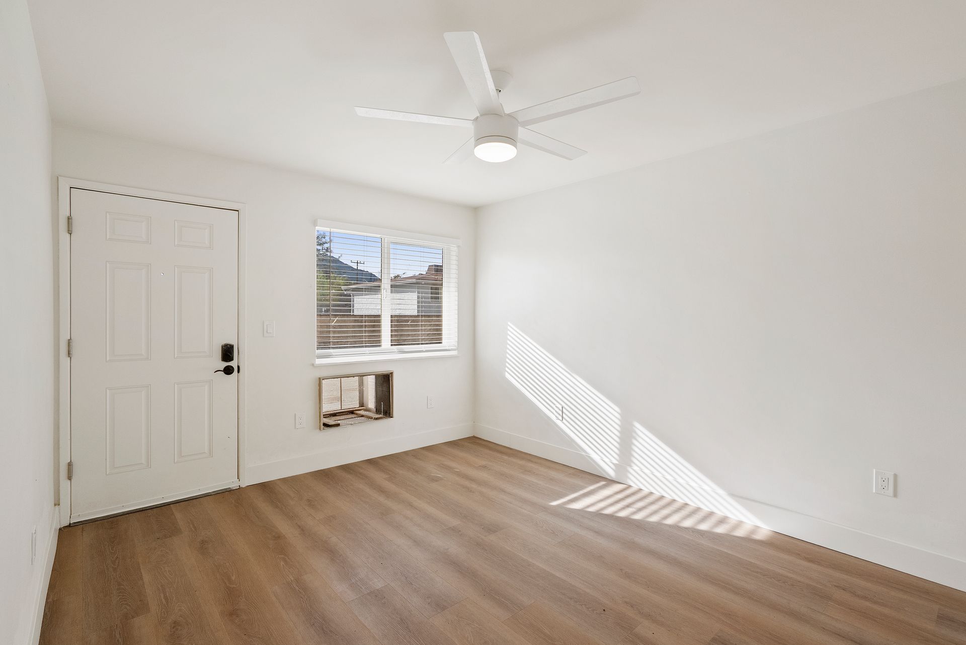 Empty, white-walled room with a door, window, ceiling fan, and wooden floor. Sunlight streams in the window.