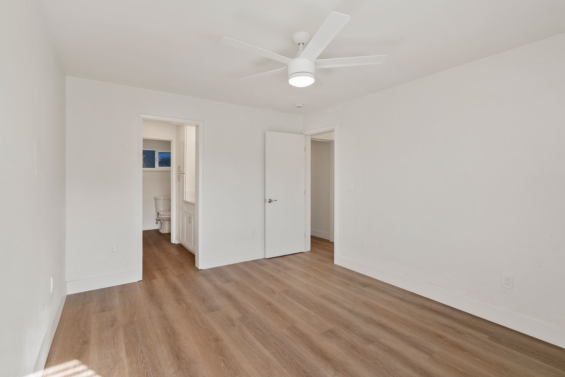 Empty bedroom with white walls, wood-look flooring, ceiling fan, and open doorways to a bathroom and another room.