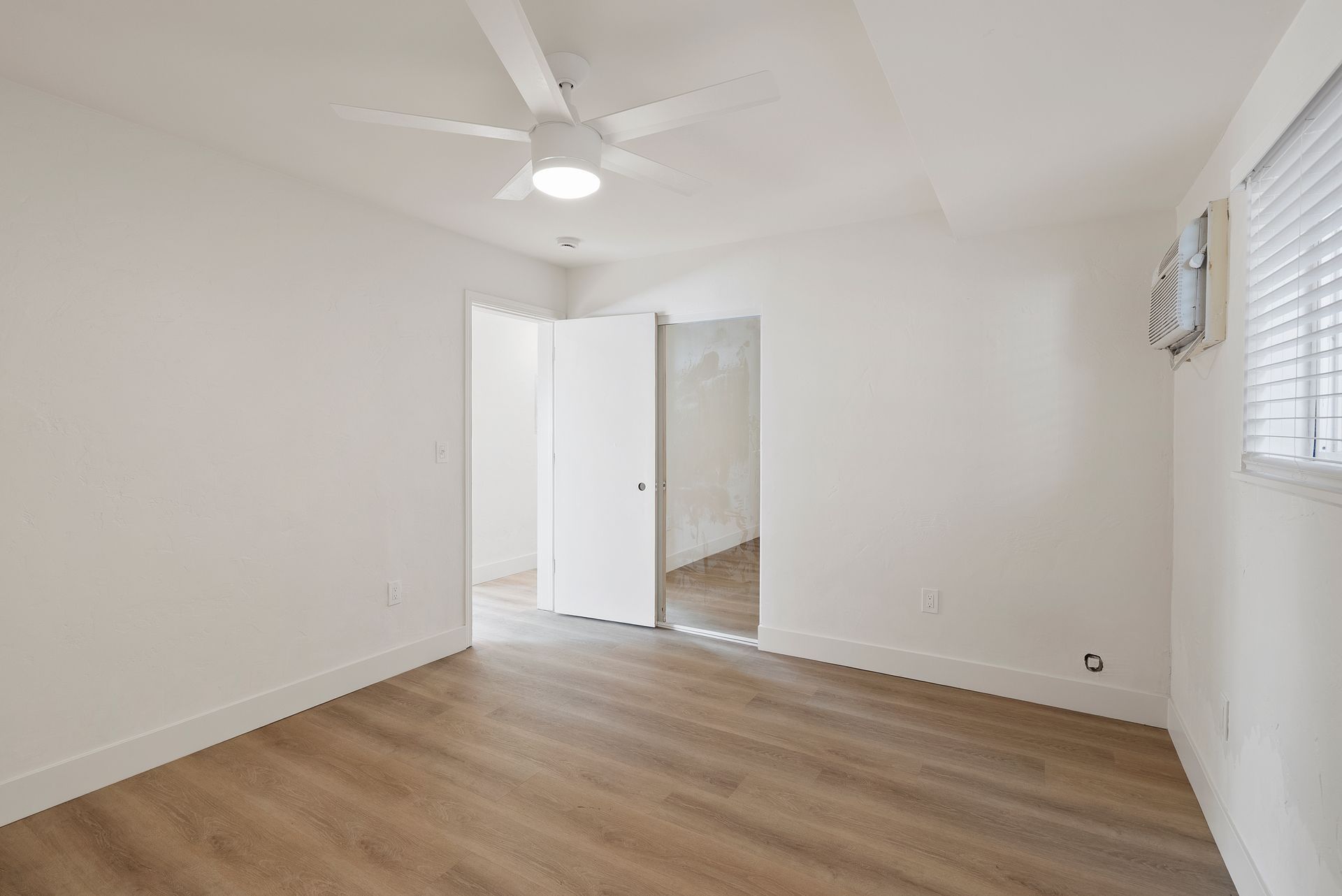 Empty white room with light wood-look flooring, door, and ceiling fan. Window with blinds on the right.