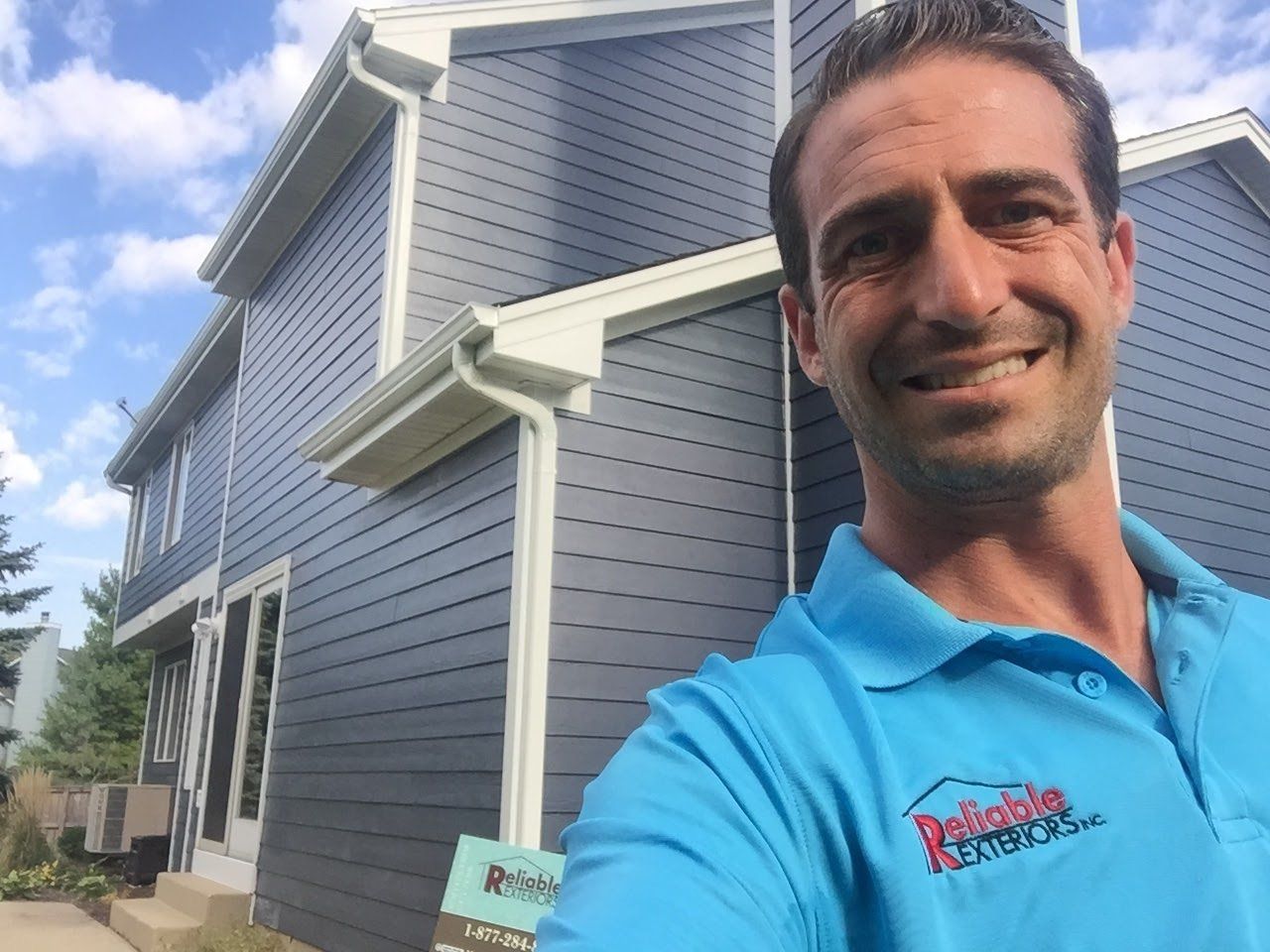 A man in a blue shirt is taking a selfie in front of a house.