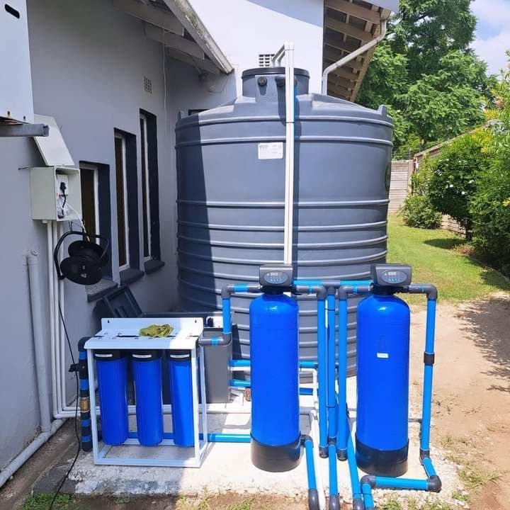 Water filtration system setup next to a gray water tank near a building. Blue pipes and tanks visible.