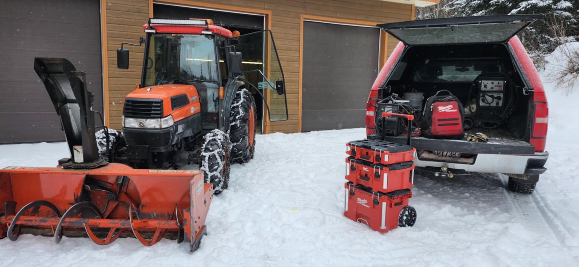 Un tracteur et une souffleuse à neige sont stationnés dans la neige devant un garage.