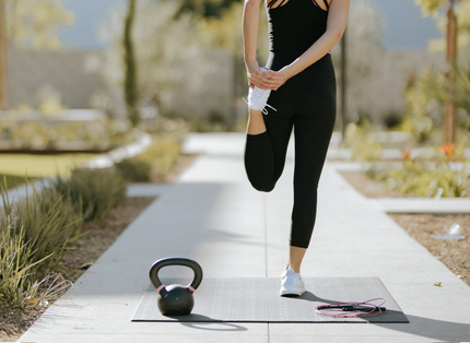 A person in black workout clothes stretches on a mat outdoors, with a kettlebell nearby.