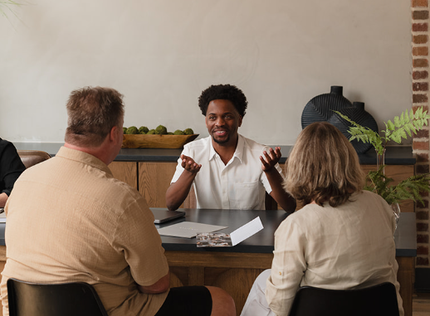 A professional meeting where three people sit around a desk, with one person gesturing while speaking to two others.