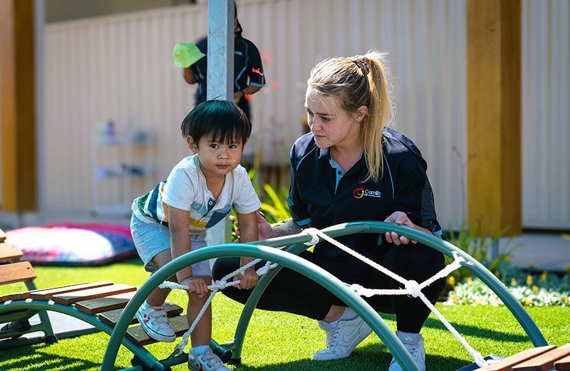Toddler playing outside on green play equipment | Carer beside toddle watching closely