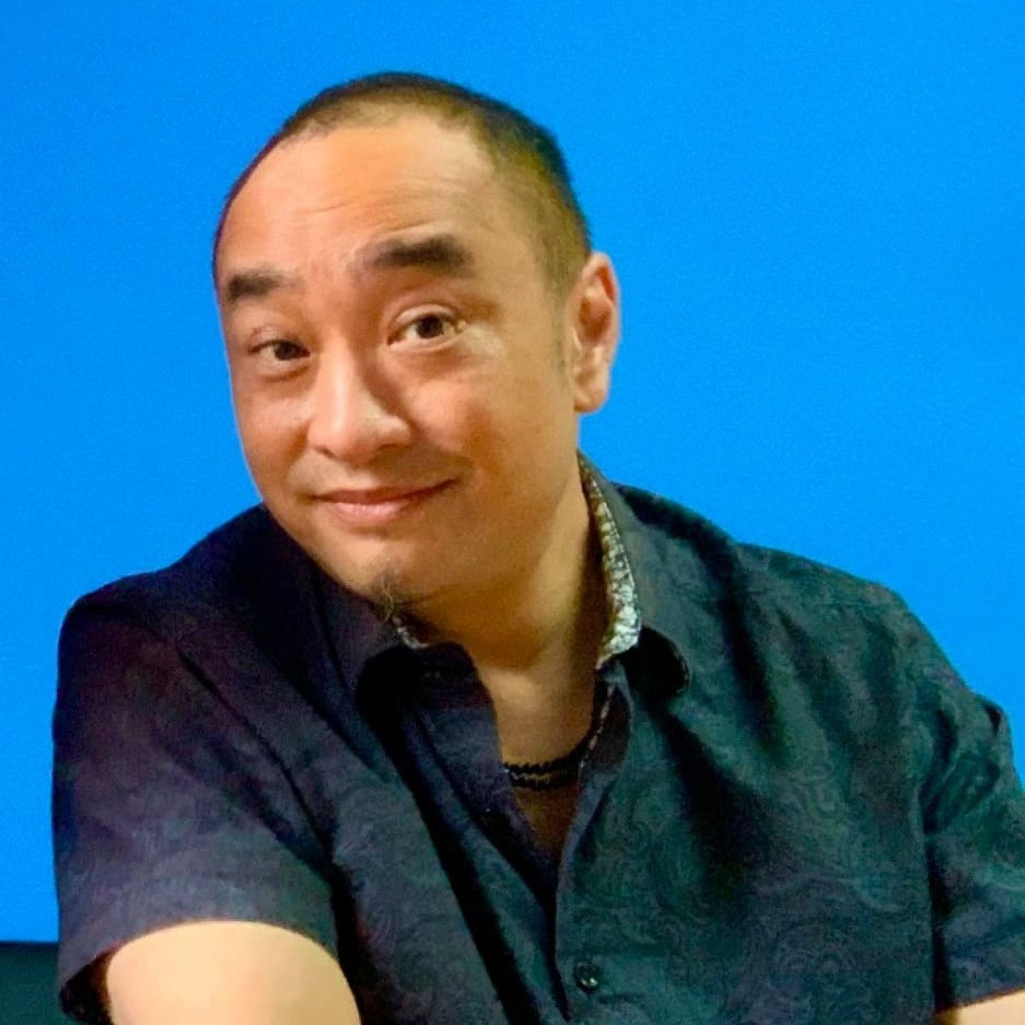 A man in a black shirt is smiling in front of a blue background.