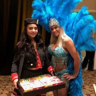 Two women in carnival costumes are standing next to each other holding a box of pizza.