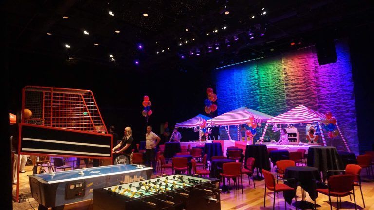 A large room with tables and chairs and a stage with a rainbow of lights.