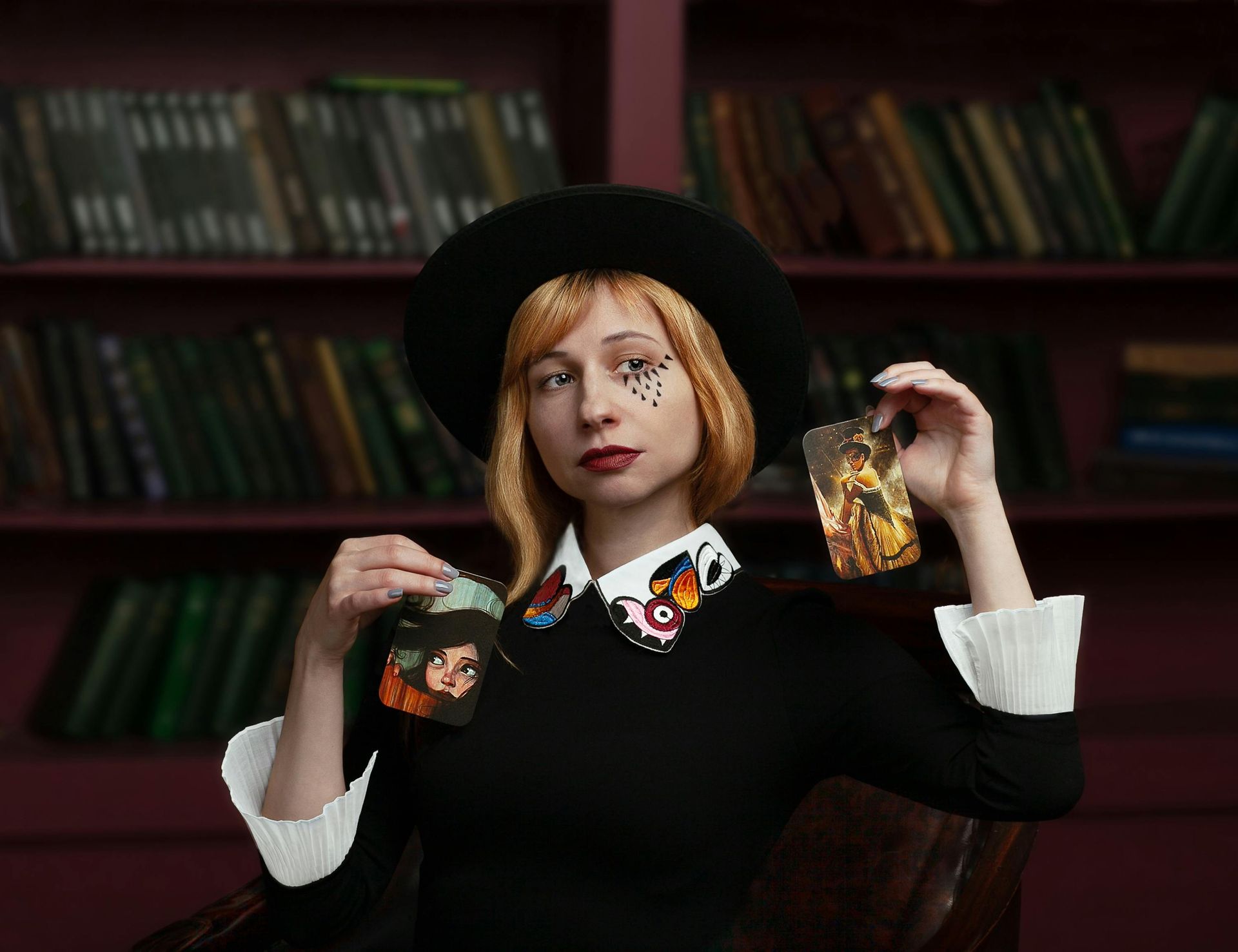 A woman in a black hat is holding a tarot card in front of a bookshelf.