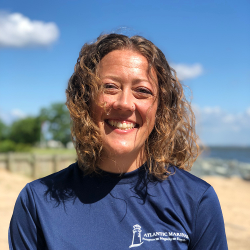 Woman smiling on a beach, wearing a blue shirt with a lighthouse logo. Sunny day, blue sky and water.