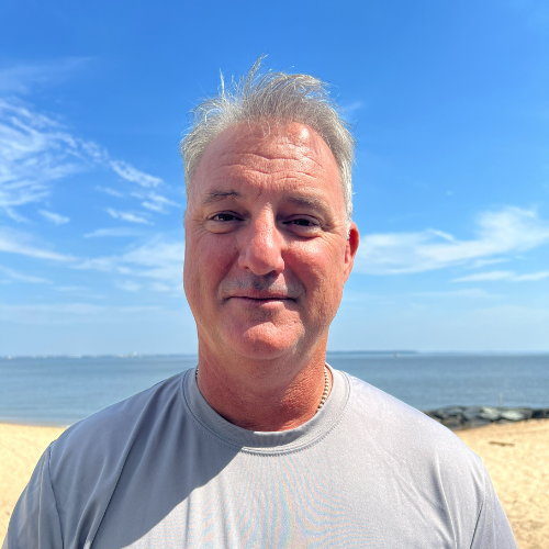 Man in gray shirt smiles at the camera on a beach with blue sky and ocean in the background.