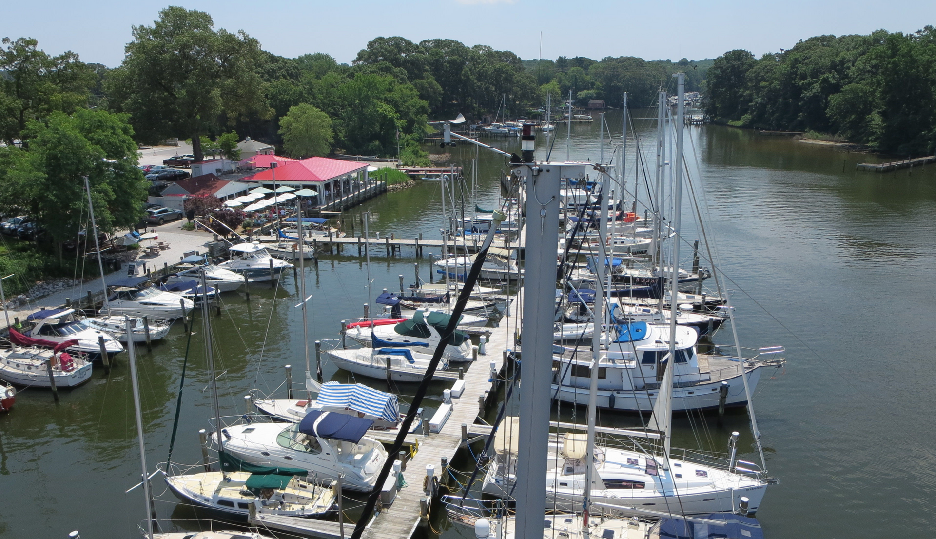 Boats docked in marina on a sunny day; red-roofed building and trees in the background.