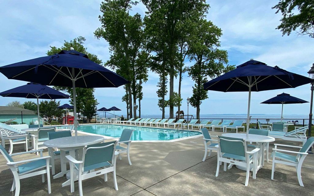 Poolside scene with blue umbrellas, tables, chairs, and a swimming pool overlooking a body of water.