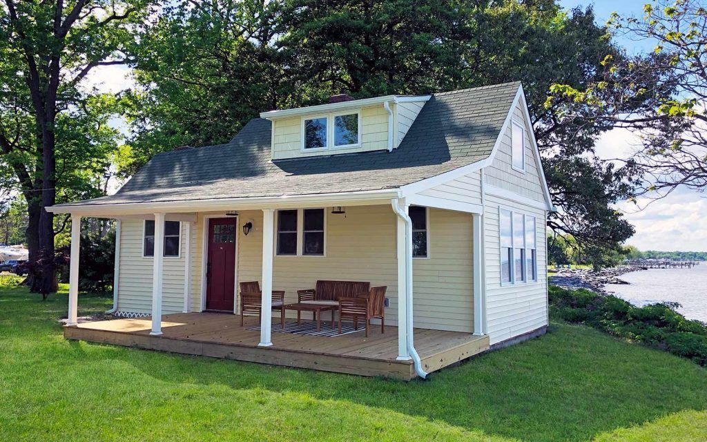 Cottage with porch overlooking water, pale yellow siding, red door, green roof, set on a grassy hill.