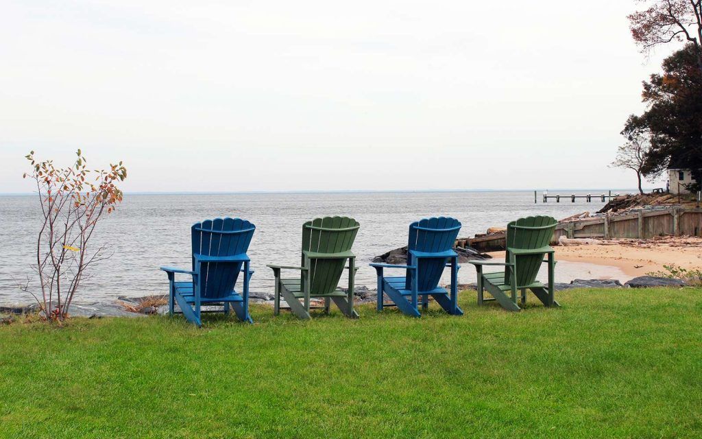 Four colorful Adirondack chairs on a grassy shore overlooking a calm body of water.
