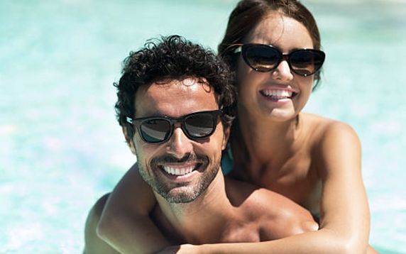 Woman hugs man from behind in a pool, both wearing sunglasses and smiling.