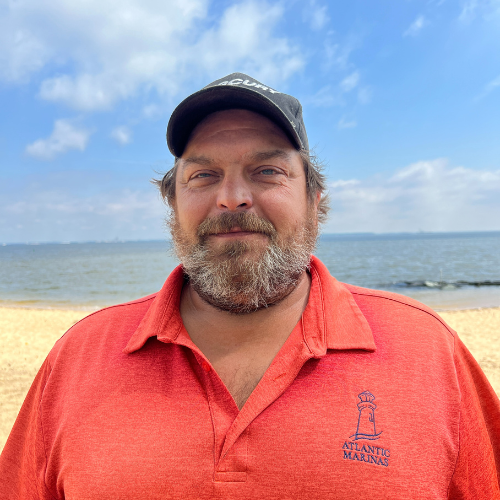 Man with a beard, wearing a baseball cap and red shirt, stands on a beach with the ocean and blue sky in the background.