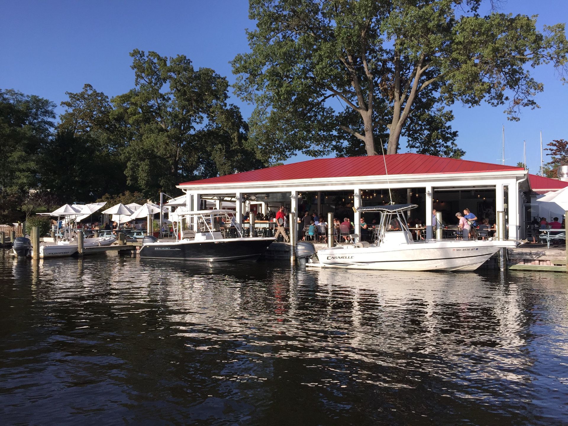 Boats docked at a waterside restaurant with outdoor seating under a red roof; trees in the background.