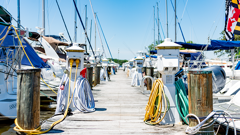 Boats docked at a wooden pier on a sunny day; yellow and green hoses hang on posts.