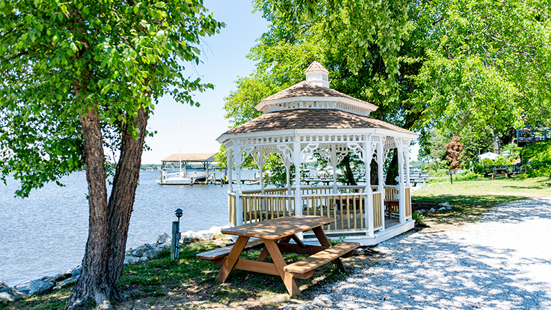White gazebo with picnic table by a lake, surrounded by trees.