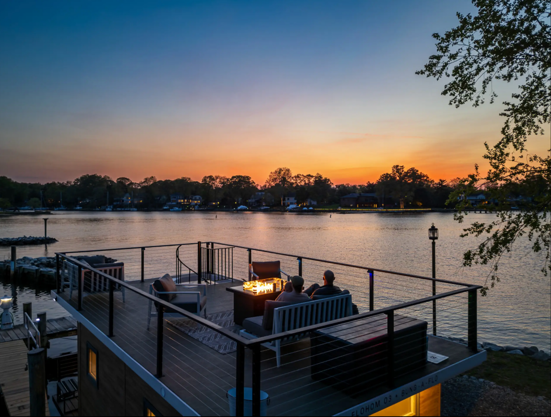 Sunset view over lake; two figures on dock, fire pit, deck, trees silhouetted, orange and blue sky.