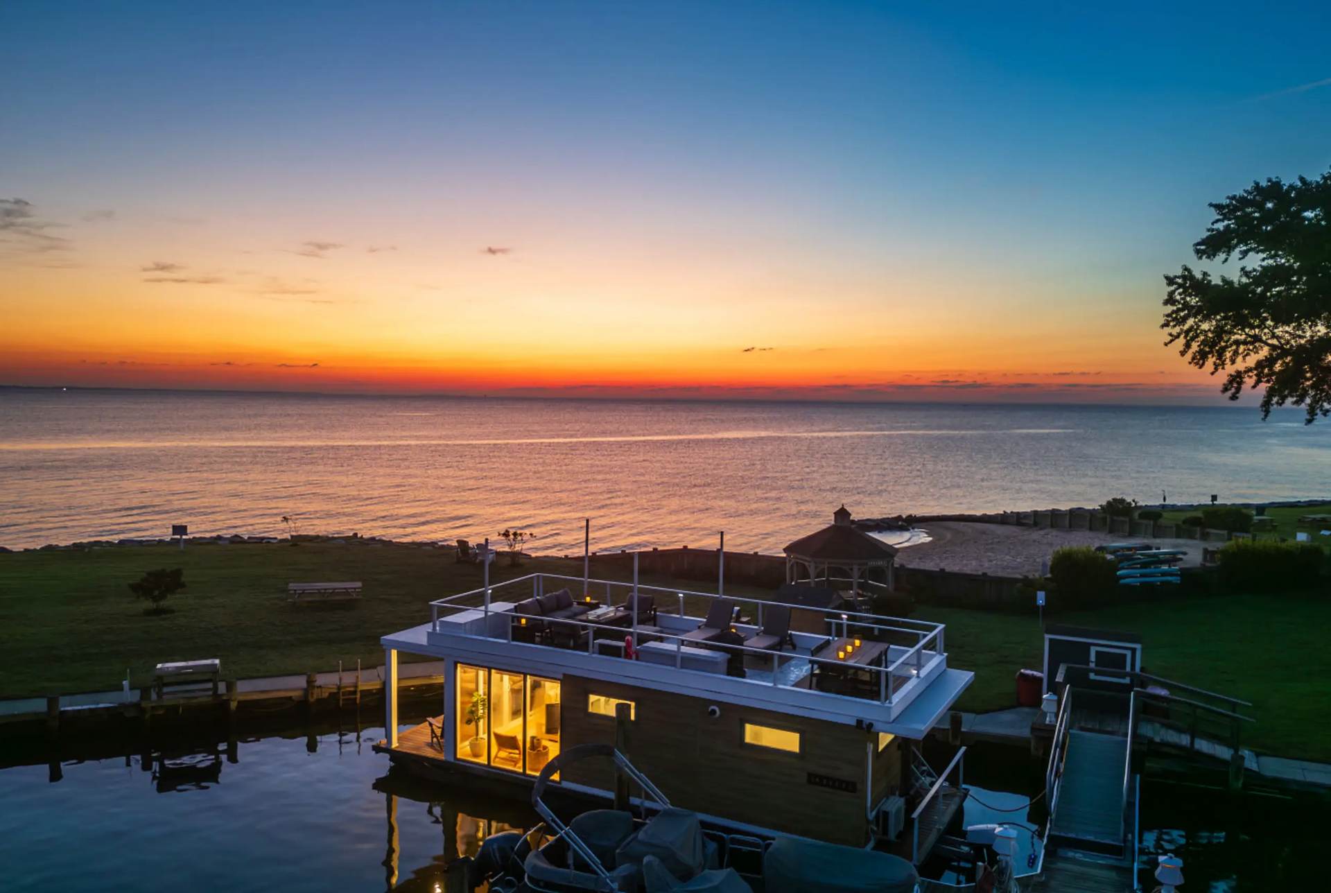 Sunset over a waterfront restaurant with outdoor seating, near a grassy lawn.