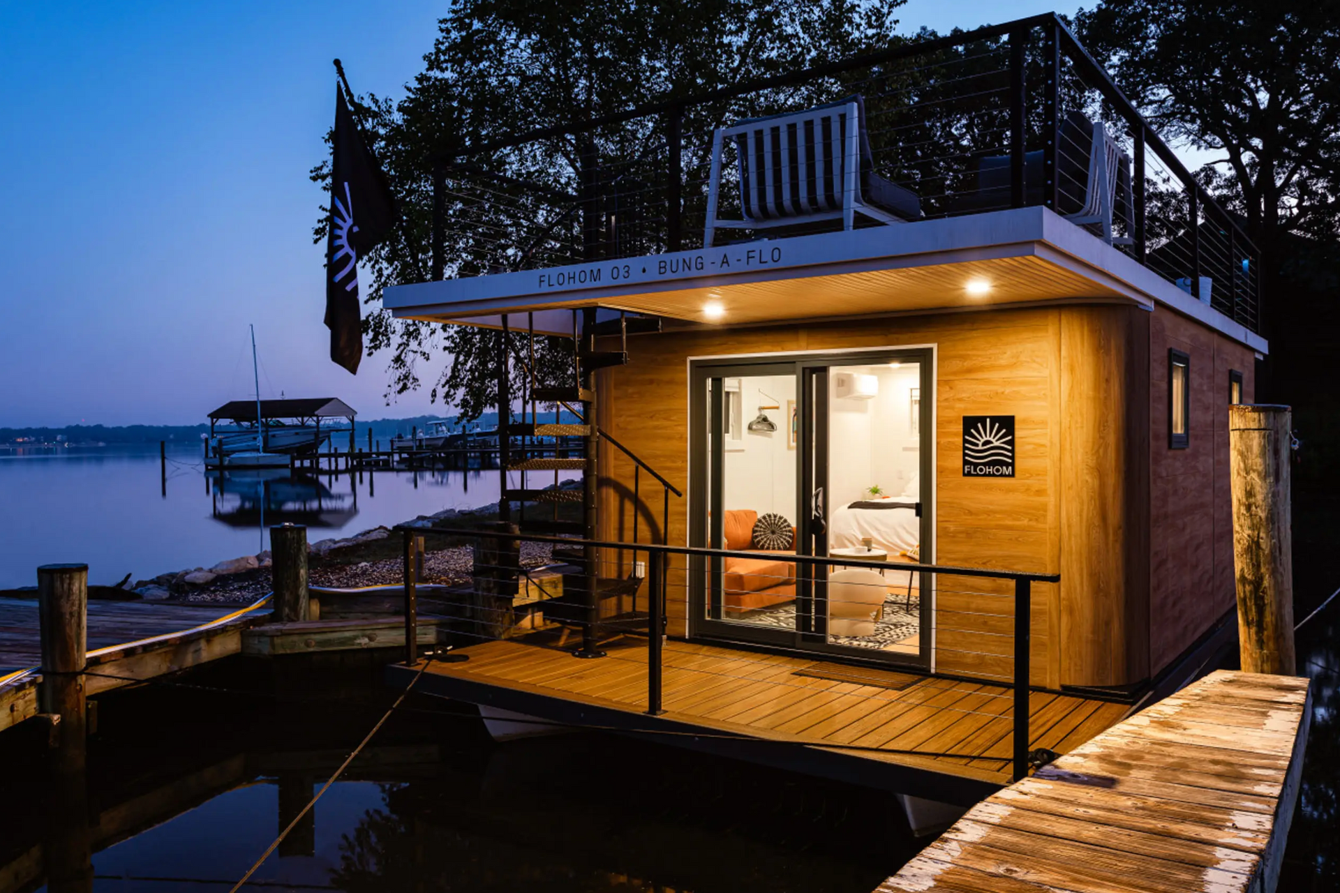 A small wooden house on a dock at dusk. Exterior lights illuminate the deck and interior.