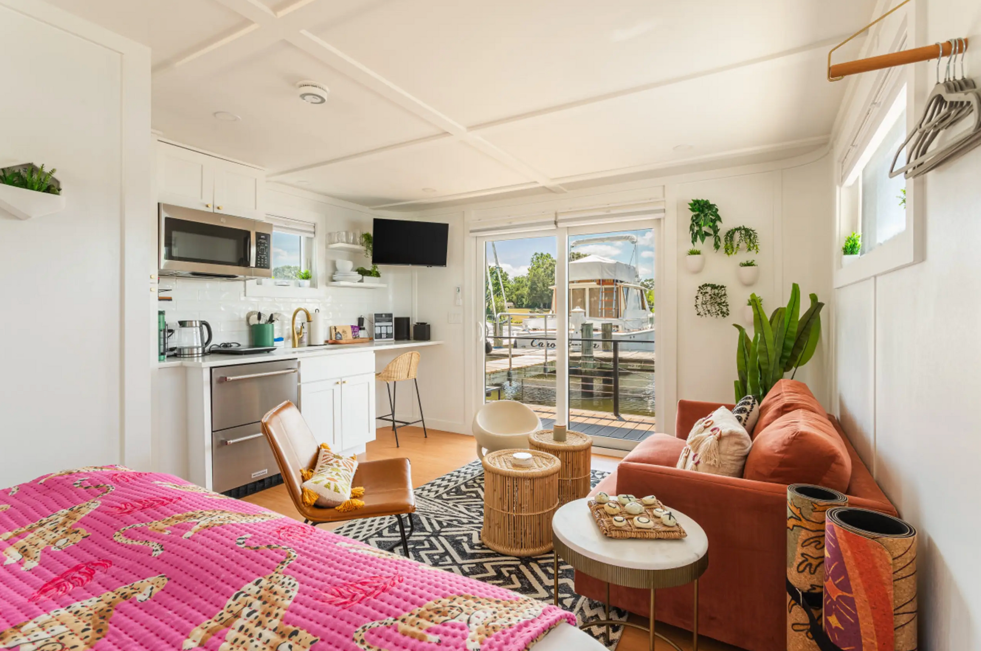 Cozy houseboat interior: kitchenette, living area with orange sofa, view of water through sliding doors, pink bedspread.