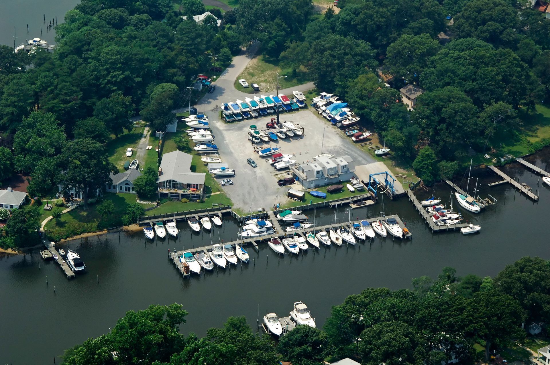 Aerial view of boats docked at a marina next to a parking lot and buildings, surrounded by trees.