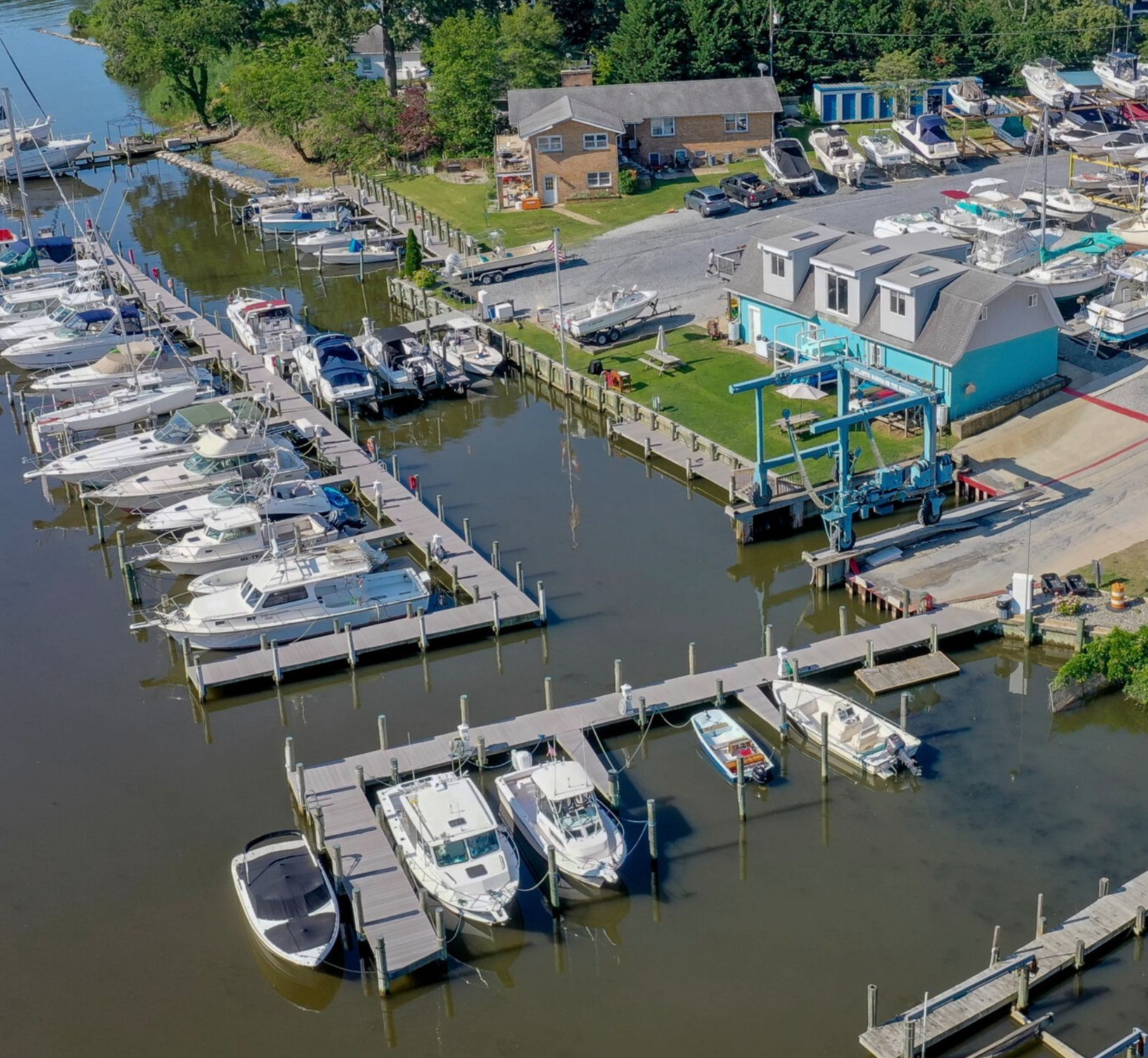 Aerial view of a marina with docks, boats, and a boat lift. Brown and blue buildings are visible.