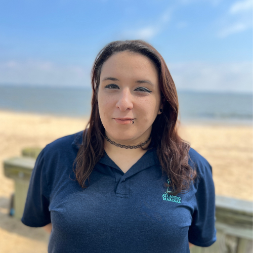 Woman with dark hair stands on a beach, wearing a blue shirt and choker.