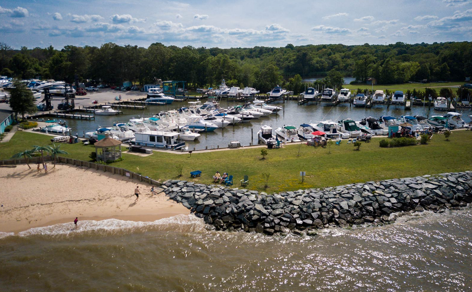 Boats docked in a harbor next to a sandy beach and grassy area with a forested background.