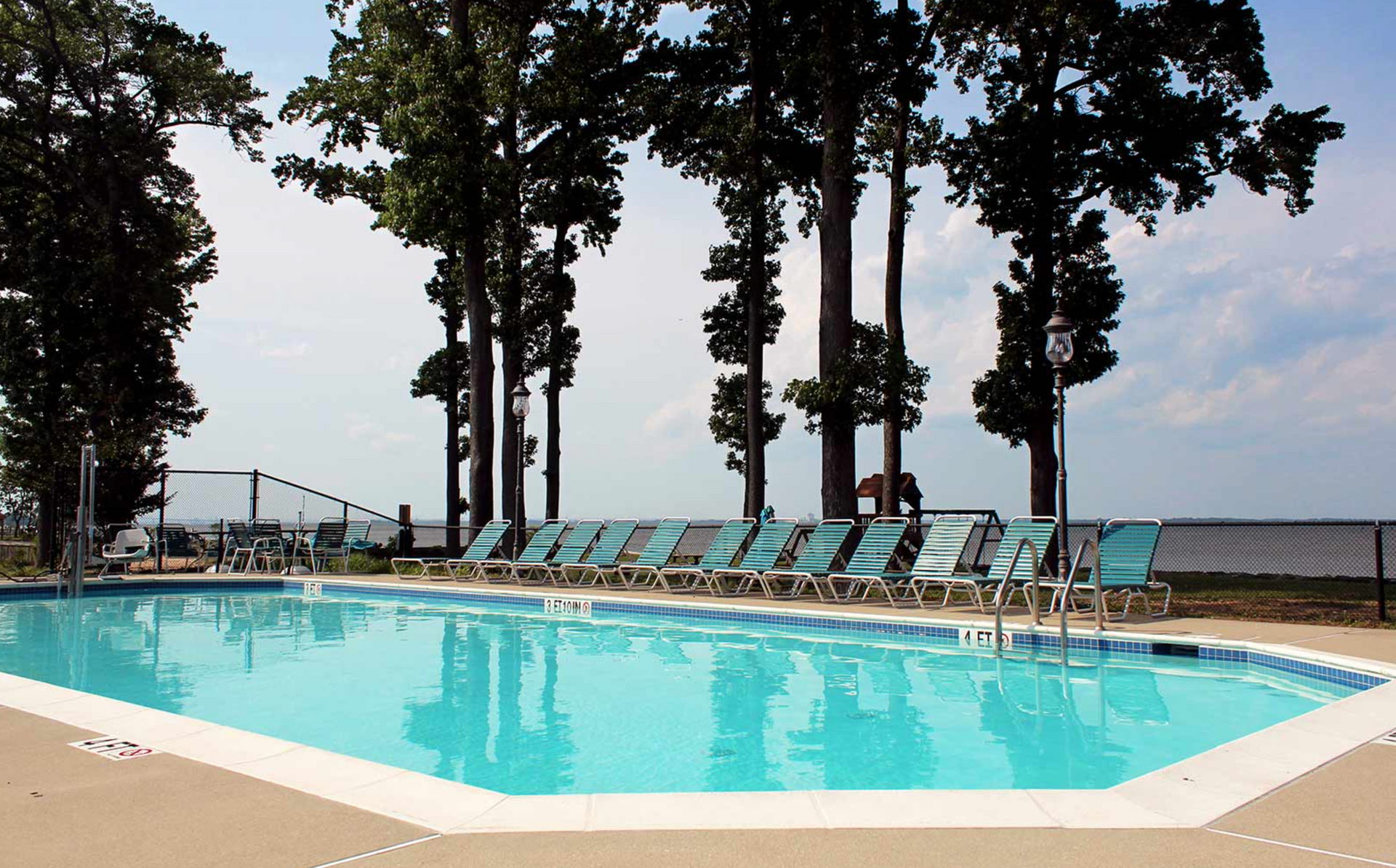 Swimming pool with turquoise water and lounge chairs, trees in the background under a cloudy sky.
