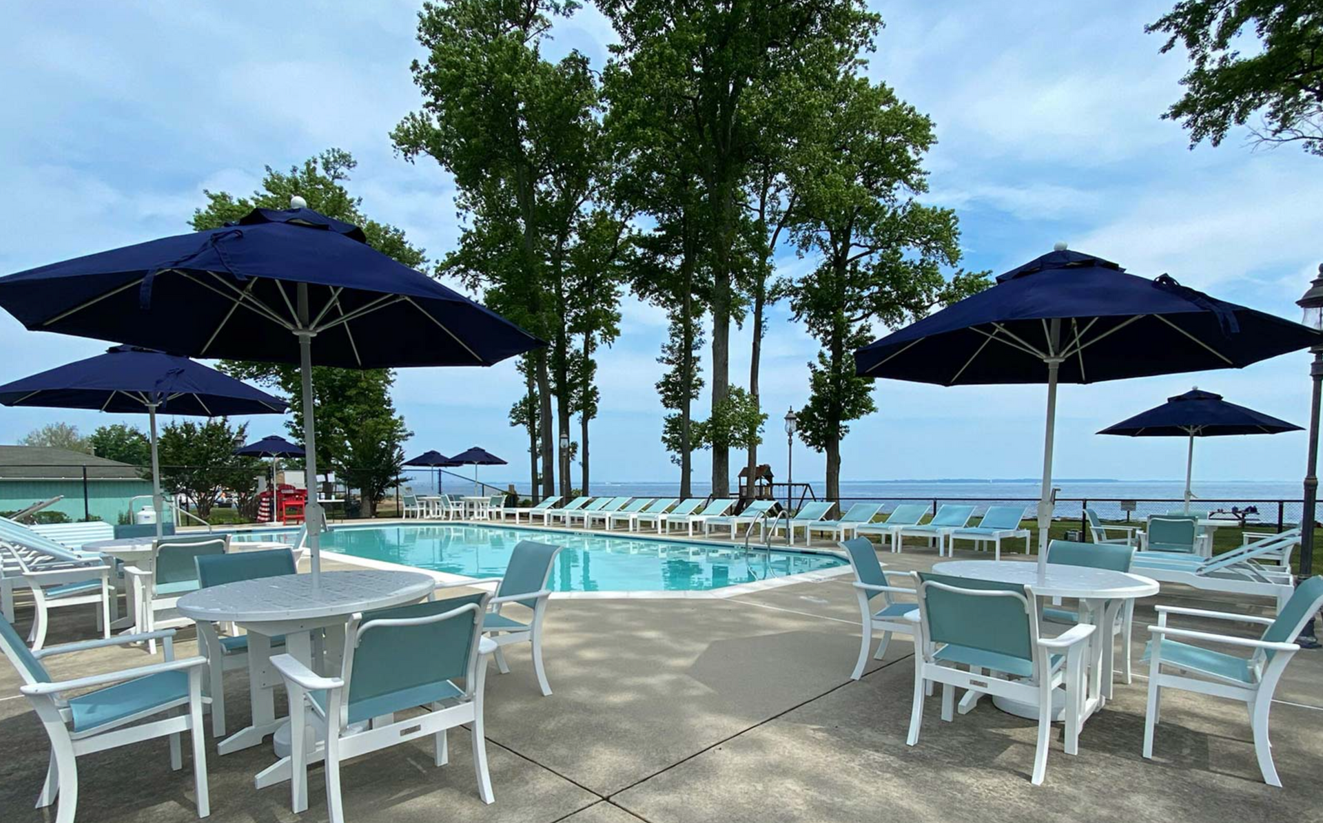Poolside scene: tables, chairs, umbrellas, and pool with lake view under a partly cloudy sky.