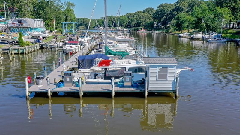 Dock with boats in a waterway, small white building, blue sky, and green trees.