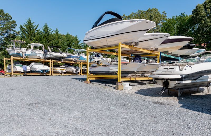 Boats stored on metal racks at an outdoor marina. Gravel lot, trees in background, sunny.