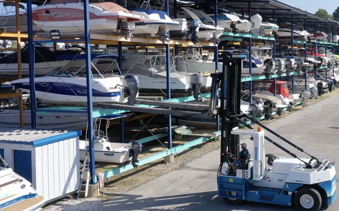 Forklift lifting boat from a multi-level boat storage rack. Boats are white, blue, and red. Outdoors.