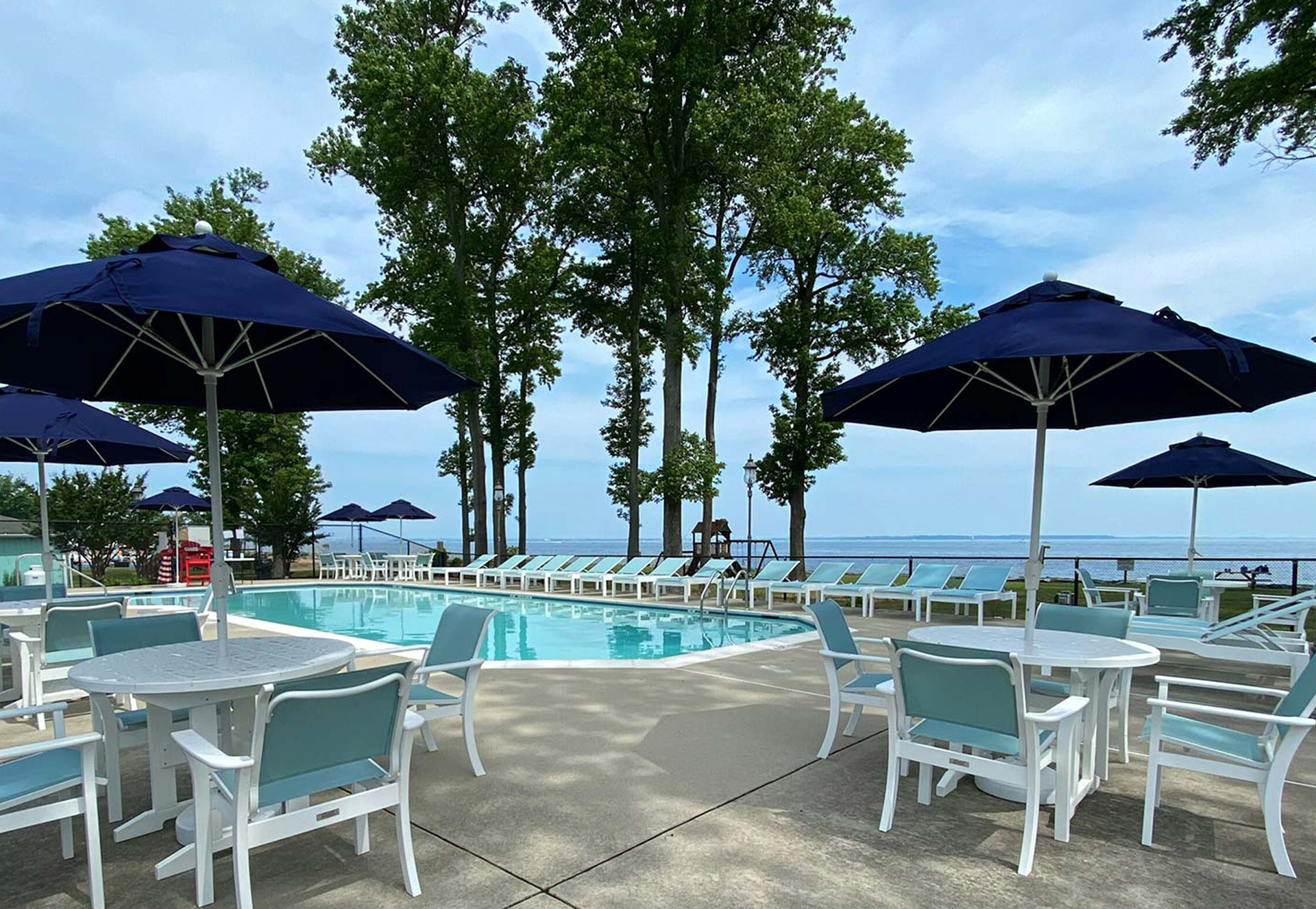 Poolside scene with tables, chairs, and umbrellas. Trees and a lake in the background under a blue sky.